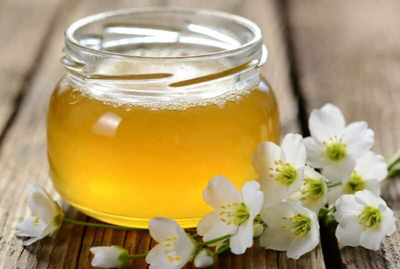 Pure golden meadowfoam honey flavor in a clear glass jar surrounded by delicate white meadowfoam flowers on rustic wood, natural sustainable Oregon honey