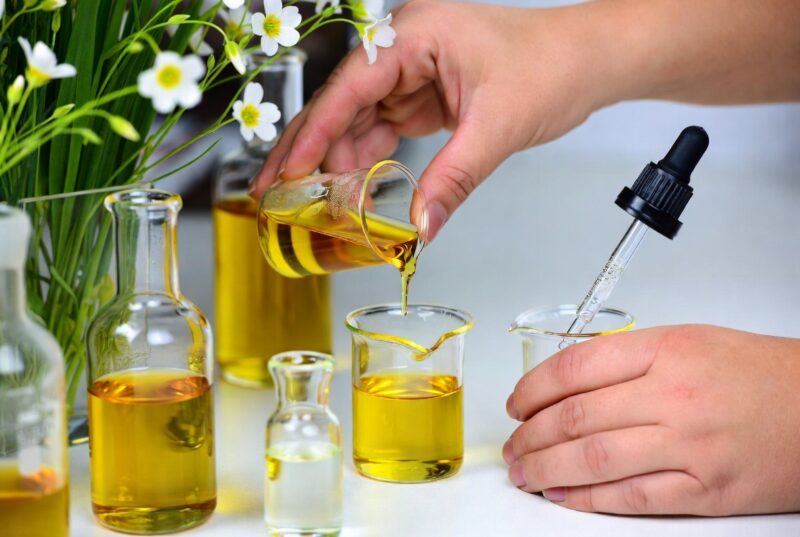 Hands carefully mixing natural seed oils in glass beakers and droppers on lab bench surrounded by meadowfoam flowers, best practices for natural skincare product formulation with botanical ingredients