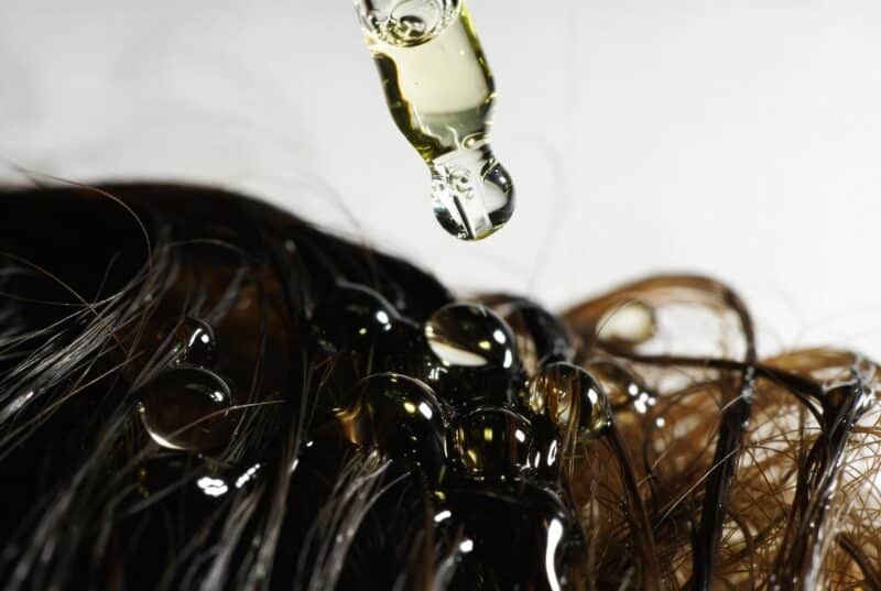 Close-up of golden meadowfoam seed oil drops from glass dropper falling onto healthy dark hair strands, antioxidant hair oil for moisture and damage repair