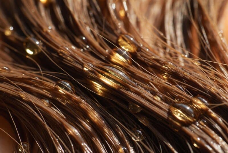 Close-up of shiny hydrated brown hair strands with golden natural carrier oil droplets, showing deep moisturizing benefits for smooth frizz-free hair