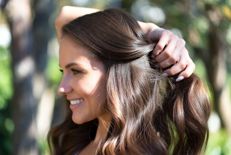 Smiling woman with long healthy wavy brown hair running her fingers through smooth frizz-free shiny strands outdoors, results from using Oregon Coriander Oil for natural hair moisture and control